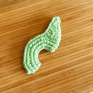 May include: A light green crocheted avocado half, with a rounded top and a curved bottom, on a wooden surface.