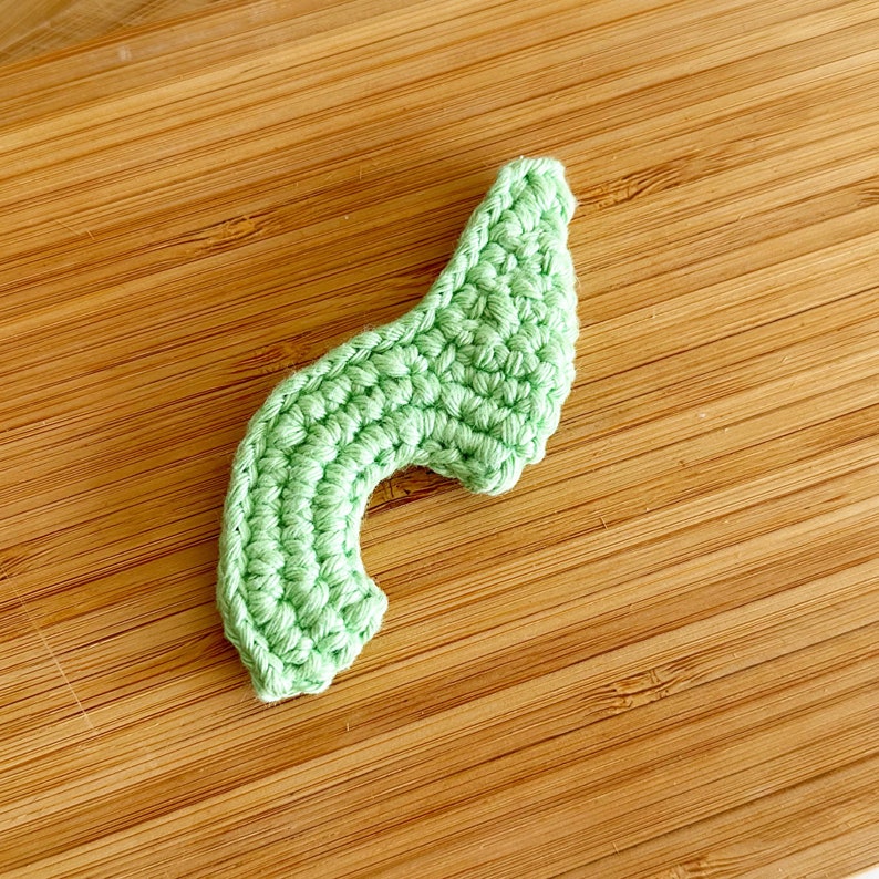 May include: A light green crocheted avocado half, with a rounded top and a curved bottom, on a wooden surface.