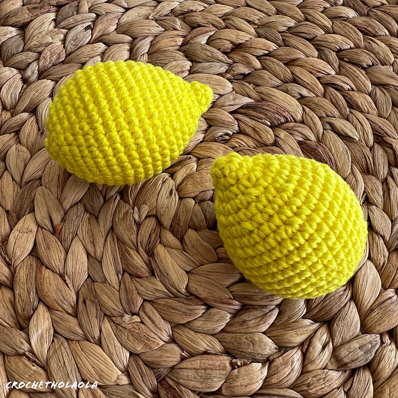 May include: Two crocheted lemon-shaped toys on a woven brown background. The lemons are yellow and have a textured surface.