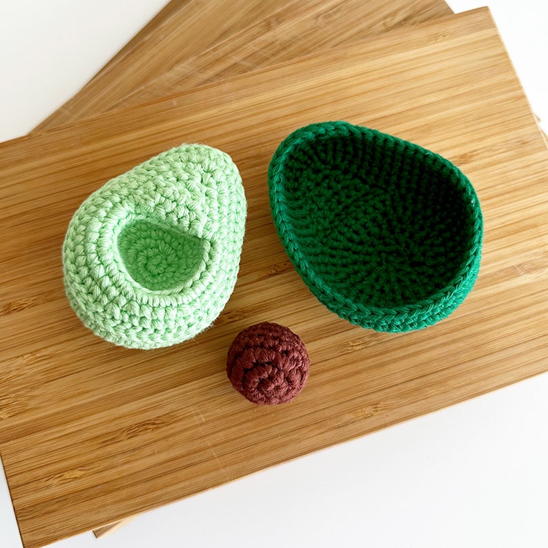 May include: A crocheted avocado set with a green avocado half, a green avocado pit, and a brown avocado seed. The set is on a wooden chopping board.