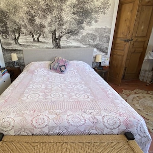 Pode incluir: Um banco retangular com assento de juta trançada em padrão espinha de peixe e pernas de madeira escura, posicionado ao pé de uma cama coberta com uma colcha de crochê branca e rosa com um motivo floral central e uma pequena almofada estampada.