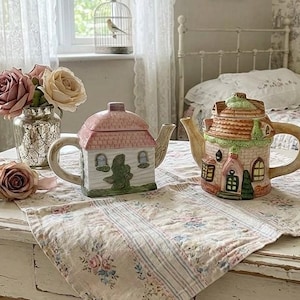 Puede incluir: Dos teteras de cerámica con forma de casitas, una con techo rosa y paredes blancas, la otra con techo multicolor y ventanas detalladas. Están colocadas sobre un paño con estampado floral junto a un jarrón de cristal con rosas rosas.