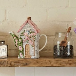 May include: A ceramic teapot shaped like a cottage with a pink roof, a brown door, and green vines with pink flowers. Beside it are a silver metal tea canister, a clear glass jar filled with loose tea leaves and a wooden scoop, and a small vase of flowers.