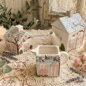 May include: A ceramic tea set featuring a house-shaped teapot, a lidded sugar bowl, and a creamer. Each piece is painted in pastel colours with a light blue roof, pink flowers, and green foliage details, designed to resemble a small cottage.