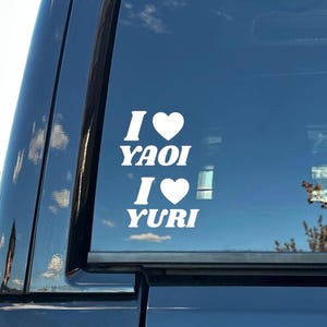 May include: White vinyl decal on a black vehicle window. The decal reads "I ♥ YAOI" and "I ♥ YURI" in a bold, sans-serif font, with a heart symbol replacing the word "love". The background shows a blue sky.