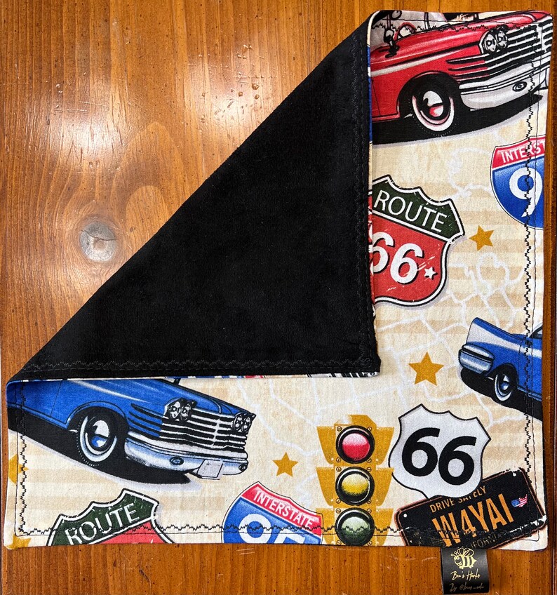 May include: A cream-colored fabric with a vintage road trip theme. It features blue and red cars, Route 66 signs, and a traffic light. The fabric has a black backing.