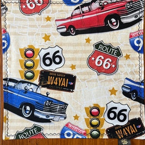 May include: A cream-colored fabric with a repeating pattern of vintage cars, traffic lights, and Route 66 signs. The signs are red, white, and blue with black lettering. The cars are red, blue, and black. The traffic lights are red, yellow, and green. The background is a cream color with a subtle pattern of stars.