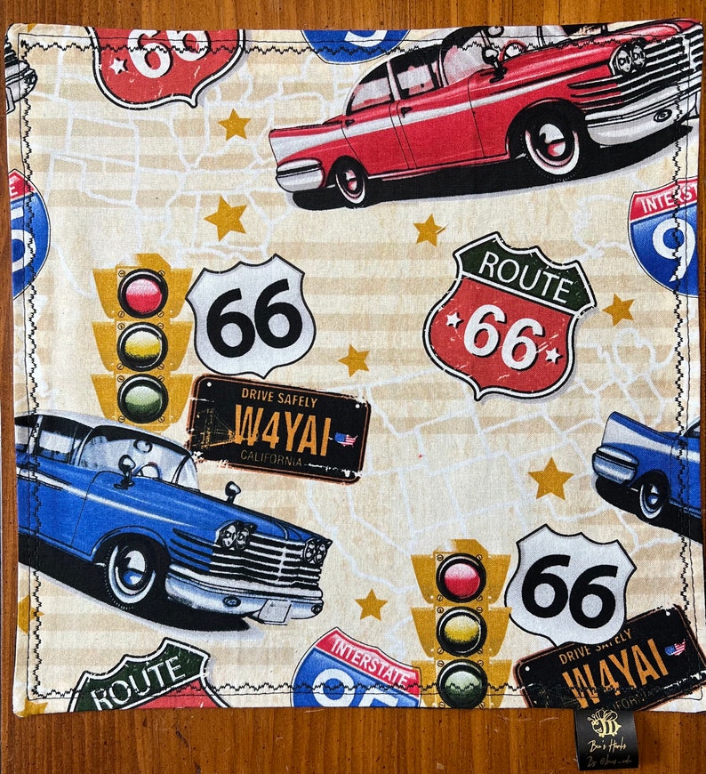 May include: A cream-colored fabric with a repeating pattern of vintage cars, traffic lights, and Route 66 signs. The signs are red, white, and blue with black lettering. The cars are red, blue, and black. The traffic lights are red, yellow, and green. The background is a cream color with a subtle pattern of stars.