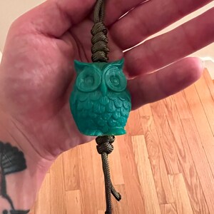 Peut inclure: Un pendentif en forme de hibou vert turquoise avec un design détaillé, suspendu à un cordon en paracorde vert.