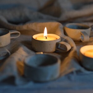 May include: A selection of small, ceramic candle holders with lit candles. The holders are cup-shaped with handles, and the soft glow of the flames illuminates the scene. The holders are arranged on a neutral-coloured fabric.