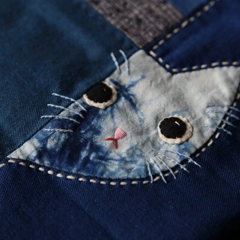 May include: Close-up of a blue fabric cat face applique. The cat's face is light blue with dark blue tie-dye effect. Features include black eyes, a pink nose, and white stitched whiskers. The applique is sewn onto a darker blue fabric.