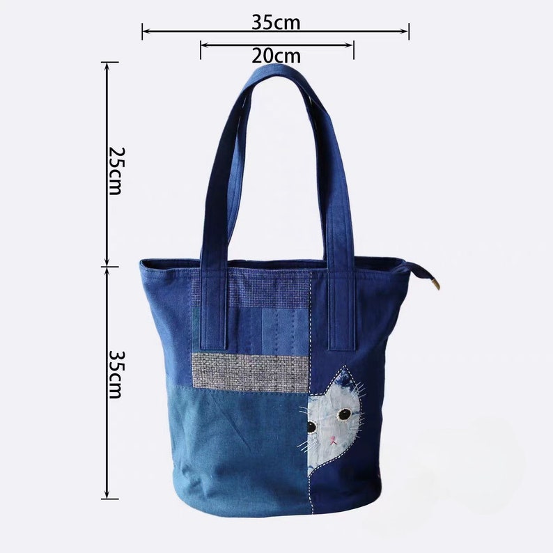 May include: A blue tote bag with a cat design. The bag is 35 cm wide, 35 cm tall, and has a handle height of 25 cm. The cat design is on the lower right side of the bag. The bag has a zipper closure.