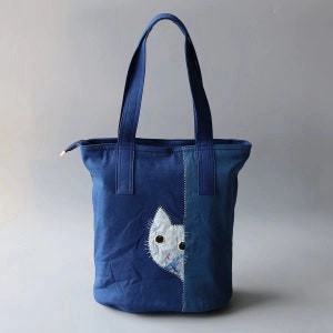 May include: A blue denim tote bag with a white cat face applique. The bag has two handles and a zipper closure. The cat applique is on the front of the bag, with black button eyes and a pink nose.
