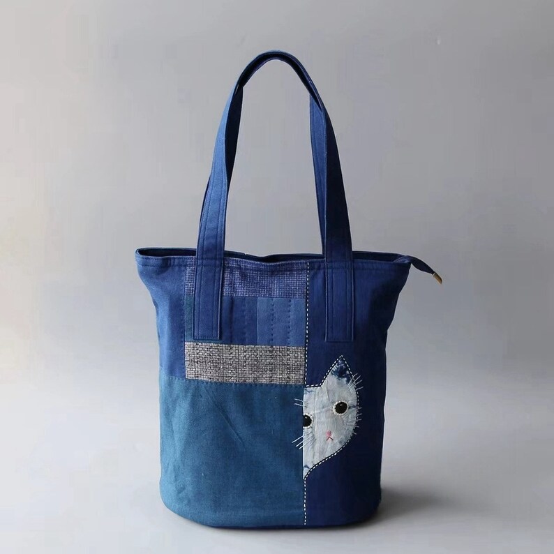 May include: A blue tote bag with a cat face applique. The bag features a patchwork design with various shades of blue fabric and a zipper closure. The cat applique is white with black eyes and a pink nose.