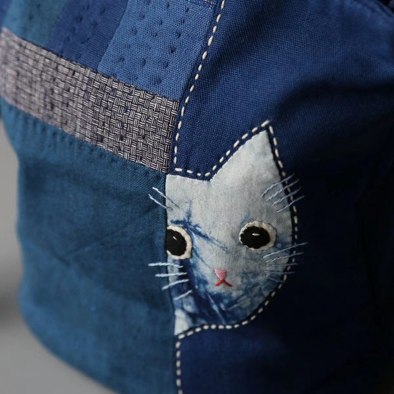 May include: Close-up of a blue fabric bag with a cat face applique. The bag is made of various shades of blue fabric, with white stitching and a light blue cat face with black eyes and a pink nose.