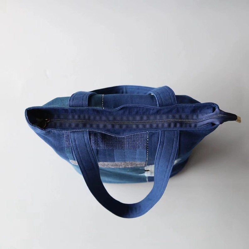 May include: A blue denim tote bag with a zipper closure and two handles. The bag features a patchwork design with various shades of blue denim. The bag is designed for carrying items.