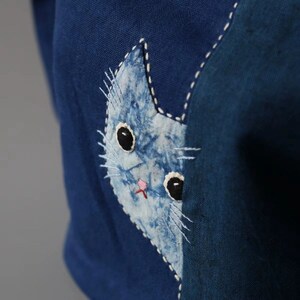May include: Close-up of a blue fabric with a cat applique. The cat is light blue with black eyes, a pink nose, and white whiskers. The cat is stitched onto the darker blue fabric with white thread.
