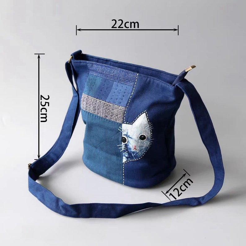 May include: A blue denim shoulder bag with a cat face applique. The bag is 25cm tall, 22cm wide, and 12cm deep. It features a patchwork design and a long strap for carrying.