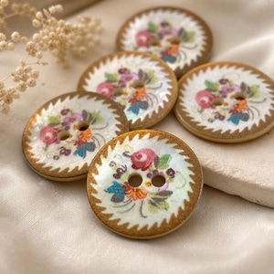 May include: Five round buttons featuring a white center with a colorful floral print of pink, orange, and blue flowers. Each button has two central holes and a metallic gold serrated edge border, creating a distinct contrast against the white surface.