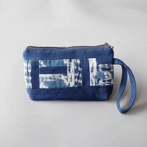 Puede incluir: Una bolsa de cremallera de mezclilla azul con un diseño de patchwork con tela de teñido anudado azul y blanco. La bolsa tiene una asa corta.