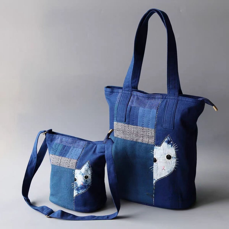 May include: Two blue denim tote bags with a cat face applique. One is a large tote bag with handles, and the other is a smaller crossbody bag with a shoulder strap. Both bags have a patchwork design.