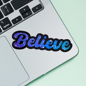 May include: A blue and purple glitter sticker with the word "Believe" in a cursive font on a silver laptop.
