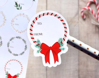 May include: A circular gift tag sticker featuring a red and green candy cane striped border, a bright red bow, and green pine needles at the bottom. The center is white with the printed text TO: and FROM: for writing recipient names.