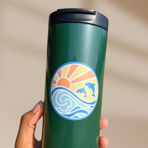 Puede incluir: Taza de viaje verde con una pegatina de un sol, olas y dos delfines en un c&iacute;rculo. La pegatina tiene un esquema de color amarillo, naranja, azul y blanco.