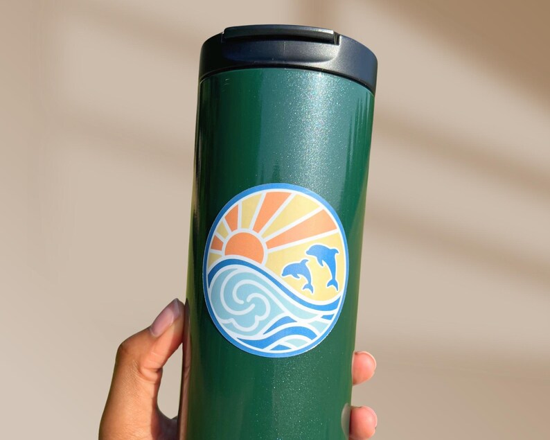 Puede incluir: Taza de viaje verde con una pegatina de un sol, olas y dos delfines en un c&iacute;rculo. La pegatina tiene un esquema de color amarillo, naranja, azul y blanco.