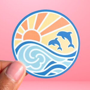 Puede incluir: Una pegatina redonda con una puesta de sol azul, blanca y naranja sobre una ola azul con dos delfines saltando fuera del agua.