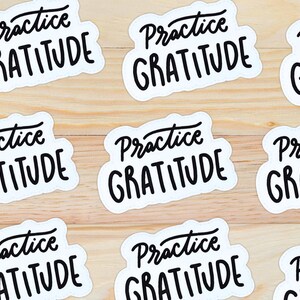 Puede incluir: Nueve pegatinas blancas con texto negro que dice "Practica la gratitud" sobre un fondo de madera claro.