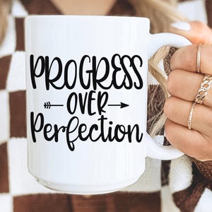 May include: White ceramic mug with the text "PROGRESS OVER PERFECTION" in black script. The mug has a large handle and is held by a person wearing rings. The background is a brown and white checkered pattern.