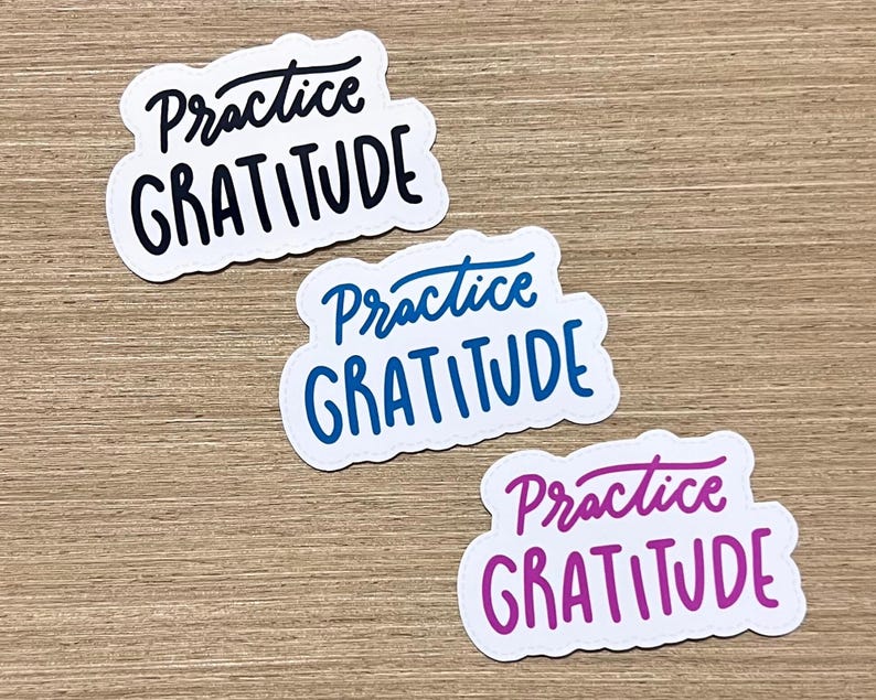 Puede incluir: Tres pegatinas blancas con bordes redondeados. Cada pegatina tiene la frase "Practice Gratitude" escrita en una fuente manuscrita. Las pegatinas son de color negro, azul y morado.