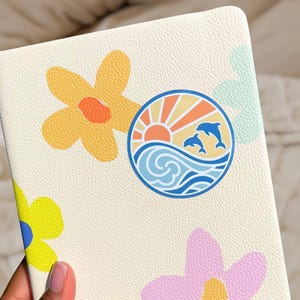 Puede incluir: Una libreta de color crema con una superficie texturizada. La libreta presenta un dise&ntilde;o colorido con un sol, olas y delfines, as&iacute; como tres flores estilizadas en amarillo, rosa y verde.