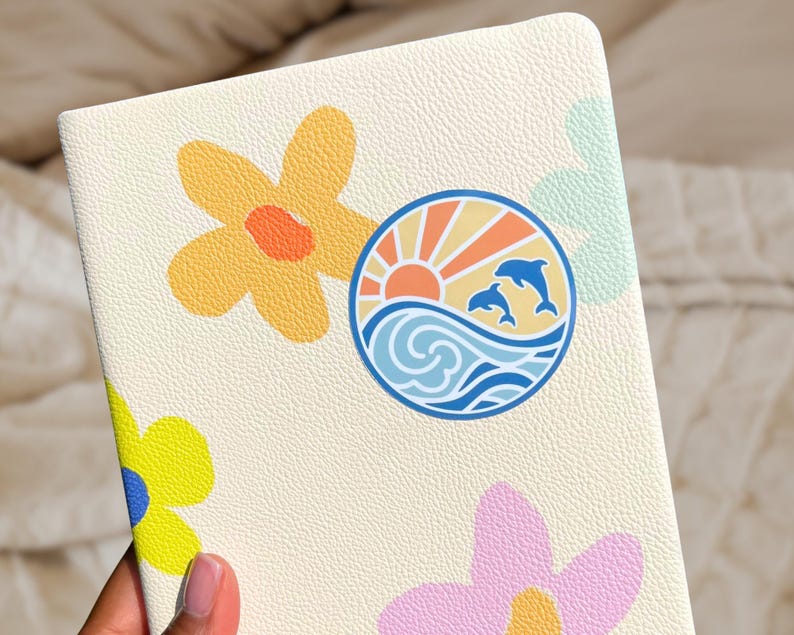 Puede incluir: Una libreta de color crema con una superficie texturizada. La libreta presenta un dise&ntilde;o colorido con un sol, olas y delfines, as&iacute; como tres flores estilizadas en amarillo, rosa y verde.