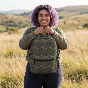 May include: A dark green backpack with a repeating pattern of tan-colored bird-like shapes. The backpack has a front pocket and a mesh side pocket. The person holding the backpack is wearing a green jacket and blue jeans.