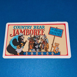 Könnte beinhalten: Ein Vintage-Aufkleber mit den Country Bear Jamboree-Charakteren von Disney. Der Aufkleber ist weiß mit einem blauen Rand und zeigt eine Cartoon-Illustration der Bären, die Musik spielen. Der Text auf dem Aufkleber lautet "Country Bear Jamboree" und "In Bear Country".