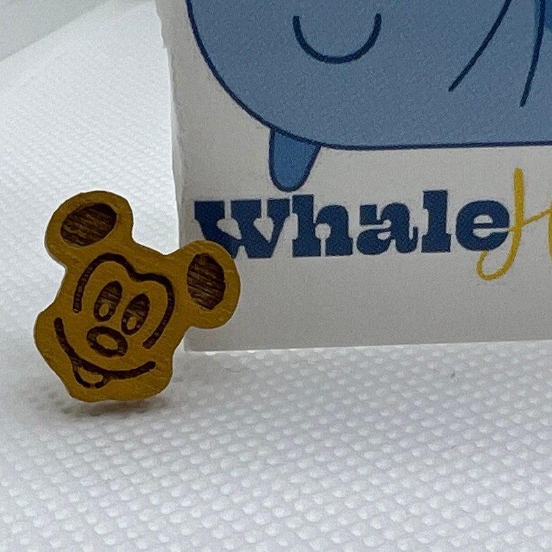 May include: A wooden Mickey Mouse head pin with a brown wood grain finish and a yellow face. The pin is smiling with a wide mouth and has two large black eyes.
