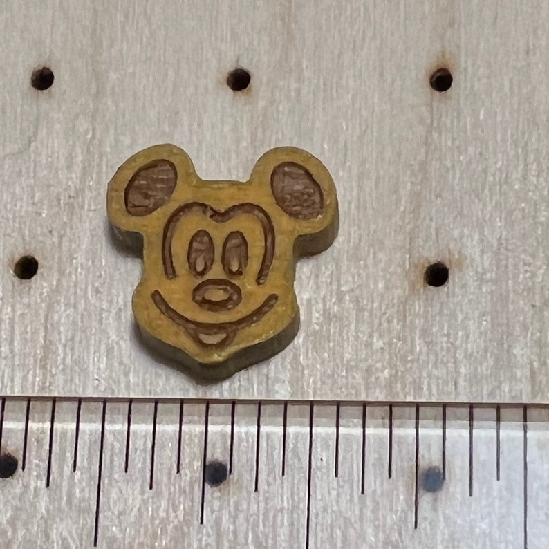 May include: A wooden Mickey Mouse head cutout with a brown outline and a yellow interior. The cutout is about 1 inch in diameter.