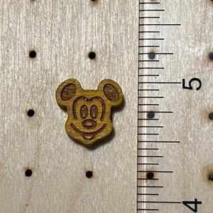 May include: A wooden button featuring a cartoon image of Mickey Mouse with a brown stain. The button is approximately 1 inch in diameter.