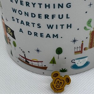 May include: A white ceramic mug with a colorful design featuring a quote that reads "Everything wonderful starts with a dream." The mug has a Mickey Mouse head pin attached to the side.