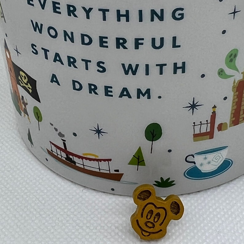 May include: A white ceramic mug with a colorful design featuring a quote that reads "Everything wonderful starts with a dream." The mug has a Mickey Mouse head pin attached to the side.