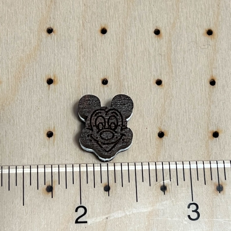 K&ouml;nnte beinhalten: Ein dunkelbrauner Holz-Knopf mit wei&szlig;em Rand, der ein Comic-Gesicht von Mickey Mouse zeigt. Der Knopf hat einen Durchmesser von etwa 2,5 cm.