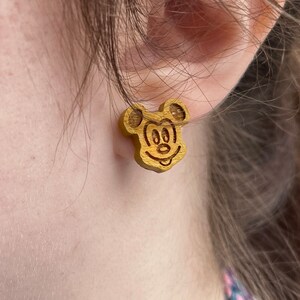May include: A pair of wooden earrings featuring a cartoon image of Mickey Mouse. The earrings are a light brown color and have a simple design.