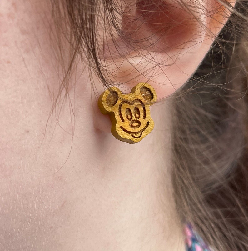 May include: A pair of wooden earrings featuring a cartoon image of Mickey Mouse. The earrings are a light brown color and have a simple design.