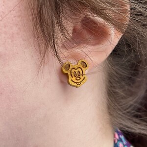 May include: A pair of wooden earrings featuring a Mickey Mouse design. The earrings are a light brown color and have a glossy finish.