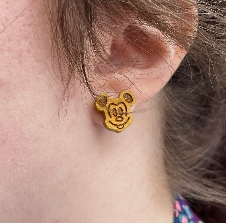 May include: A pair of wooden earrings featuring a Mickey Mouse design. The earrings are a light brown color and have a glossy finish.
