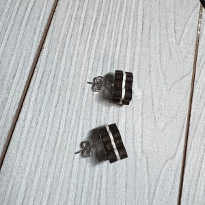 K&ouml;nnte beinhalten: Ein Paar braune und wei&szlig;e Holzohrringe mit silbernen Stiften. Die Ohrringe sind in Form von kleinen Quadraten mit wei&szlig;en Streifen.