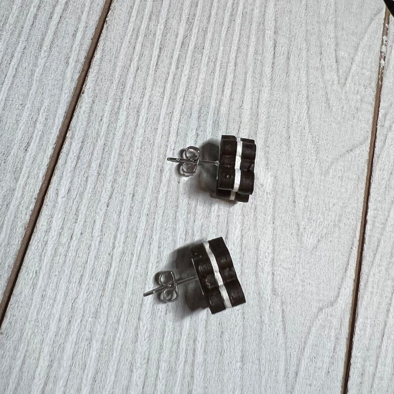 K&ouml;nnte beinhalten: Ein Paar braune und wei&szlig;e Holzohrringe mit silbernen Stiften. Die Ohrringe sind in Form von kleinen Quadraten mit wei&szlig;en Streifen.