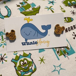 May include: A pair of wooden earrings featuring a cartoon mouse head design. The earrings are brown with a yellow outline and are displayed on a white card with a blue whale illustration and the text "Whale Honey TM".
